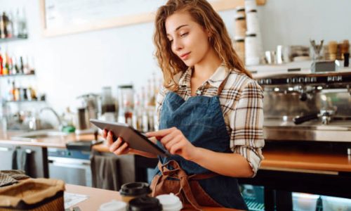 A young woman in an apron with a digital tablet at the bar counter of a coffee shop takes an order.